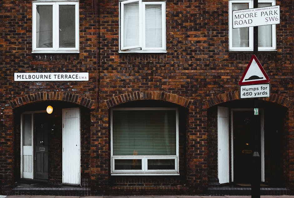 The image shows a section of a red-brick residential building with two white-framed windows on the upper floor and two on the ground floor, each with closed white blinds. The front facade features arc