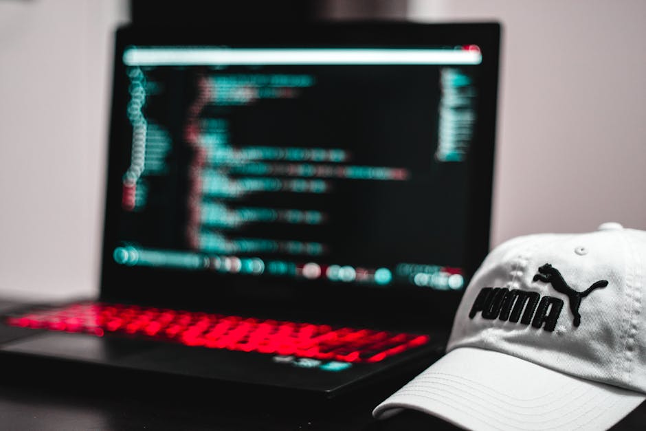 Close-up of a person's hands typing on a laptop keyboard placed on a dark table. The laptop screen displays lines of code in colorful syntax highlighting against a black background. The environment ap